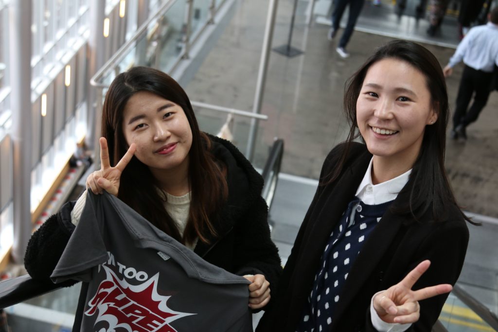 Two smiling students on an escalator, each making a peace sign, with one holding a T-shirt printed with a comic-style design