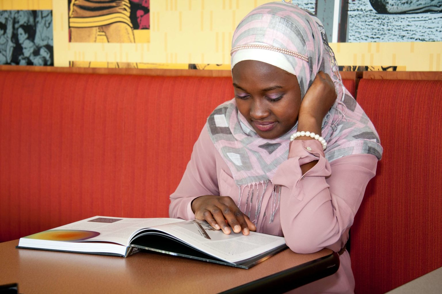 Young woman wearing a pink and gray hijab reading a book at a table in a cafe or study area with a red cushioned bench